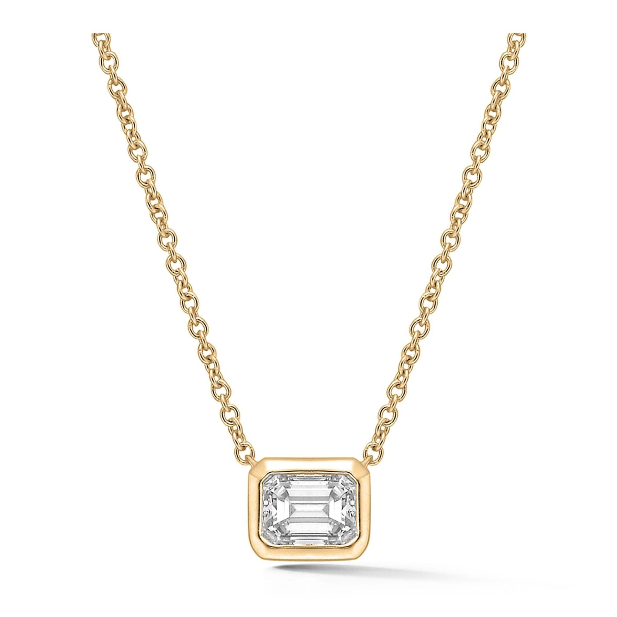 The Bezel Set Emerald Cut Diamond Solitaire Pendant in Yellow Gold features a striking rectangular emerald cut diamond elegantly encased in a polished 18K yellow gold bezel, suspended from a classic chain necklace.
