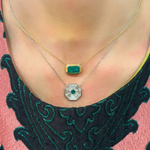 A person wearing two necklaces: the East-West Double Bezel Set Pendant with Emerald Cut Emerald in Yellow Gold, and a silver chain with a geometric silver pendant set with a small round green stone, against a patterned dark green and pink top.