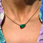 A close-up of a person wearing the East-West Pear Shape Emerald Pendant in Yellow Gold, set against bare skin and a partially visible floral-patterned garment.