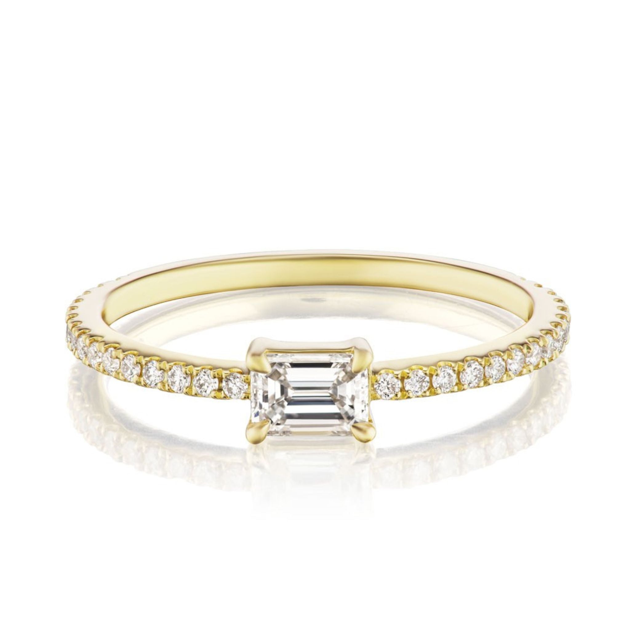 The East-West Set Emerald Cut Diamond Ring with Pavé Band in Yellow Gold showcases a rectangular emerald-cut diamond center and a slim band adorned with pavé-set round diamonds.
