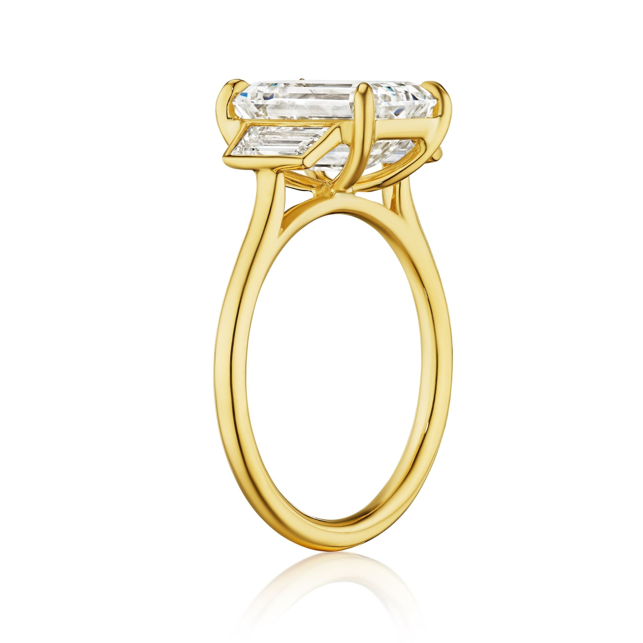 The Emerald Cut Diamond Engagement Ring with Trapezoid Side Stones features a large emerald cut diamond in 18k yellow gold, secured by four prongs, and is shown from the side on a white background.