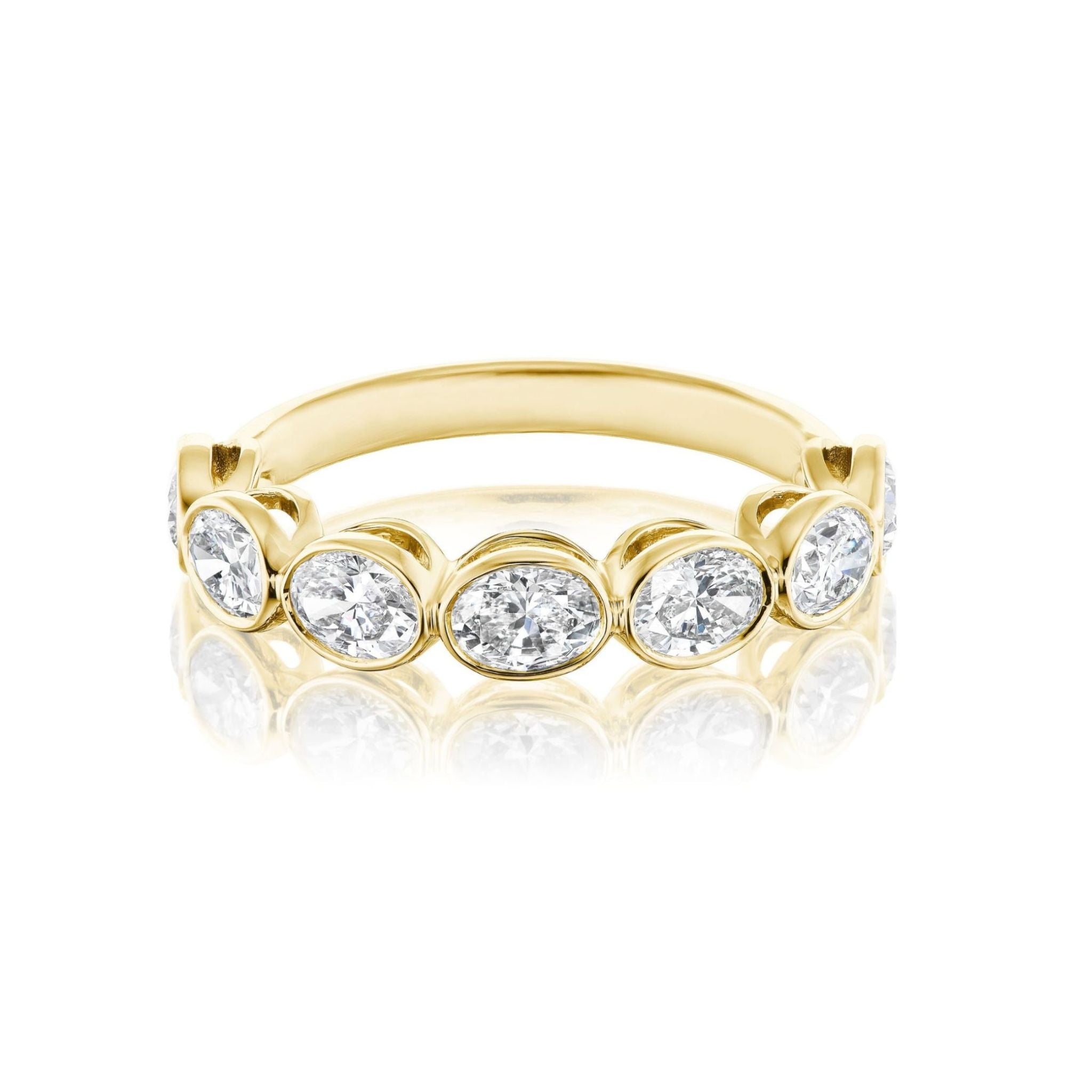 Introducing the Chasing Ovals Bezel Set Diamond Half Eternity Band—a breathtaking 18k yellow gold ring showcasing a row of oval-cut white diamonds, each nestled in its own delicate bezel. Presented on a crisp white background with a gentle reflection, this piece is exquisitely designed to elevate any bridal ring collection with timeless elegance.