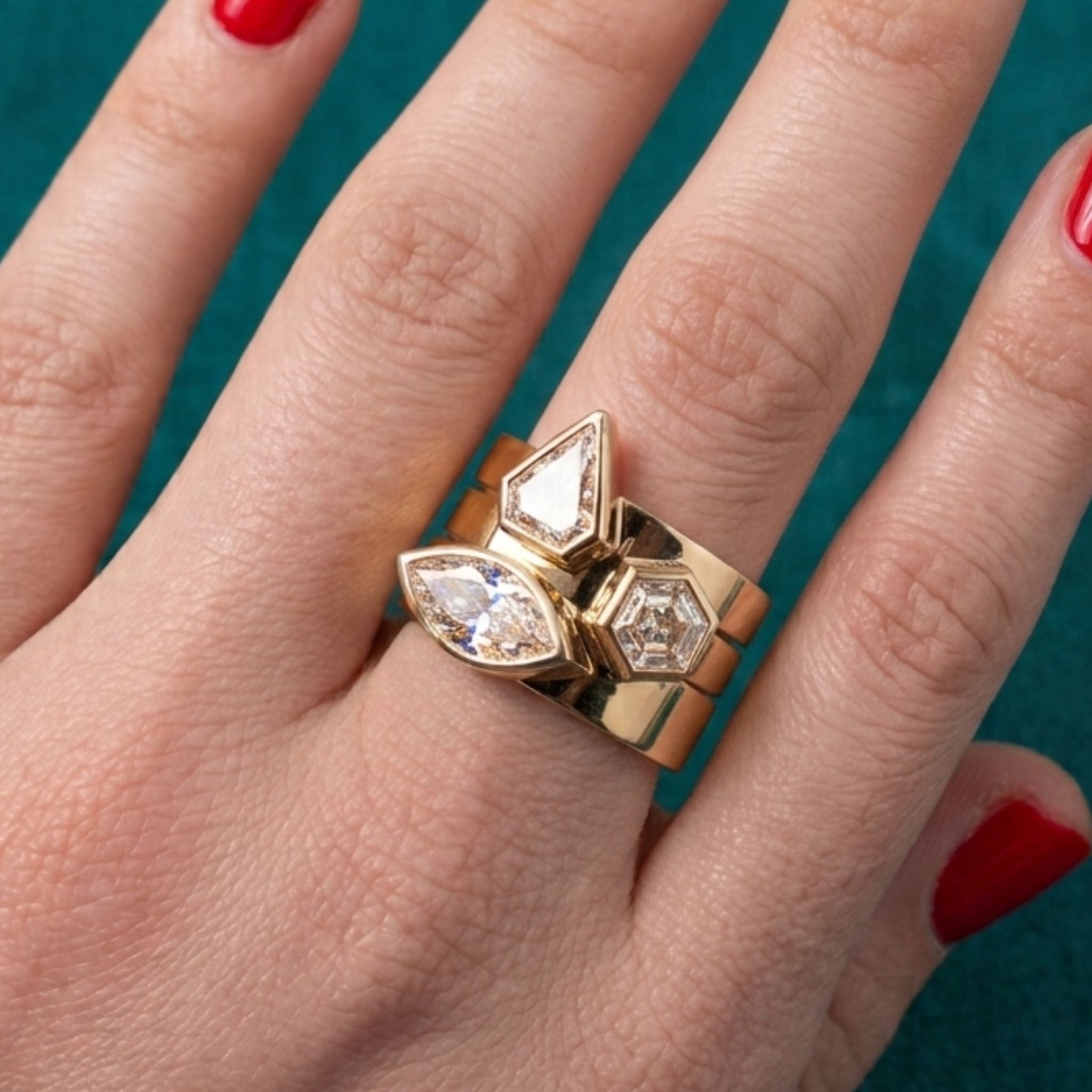 A hand with red nail polish wears three Bezel Set Mixed Shape Diamond Thick Bands in Yellow Gold, each featuring uniquely shaped clear diamonds, against a teal fabric background.