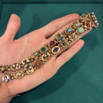 A hand displays the Colored Stone and Diamond Bracelet, crafted in 14K yellow gold with ornate links, colored stones, diamonds, intricate details, and a small painted portrait at one end. The background is green.