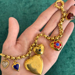 A hand displays the Gold, Colored Stone and Diamond Charm Bracelet with puffy heart charms in gold, blue, red, and jeweled designs against a green fabric background.