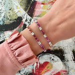A person in a pink, ruffled sleeve shows off two jeweled bracelets—one is the Multicolor Emerald Cut Sapphire Tennis Bracelet—featuring pink, red, and clear stones, set against a colorful abstract painted background.