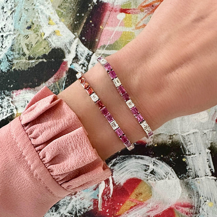 A person in a pink, ruffled sleeve shows off two jeweled bracelets—one is the Multicolor Emerald Cut Sapphire Tennis Bracelet—featuring pink, red, and clear stones, set against a colorful abstract painted background.