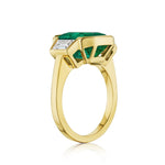 The Half Bezel Set Emerald Cut Emerald Engagement Ring features trapezoid diamond side stones and a polished 18k yellow gold band, shown at an angle to highlight its modern geometric setting.