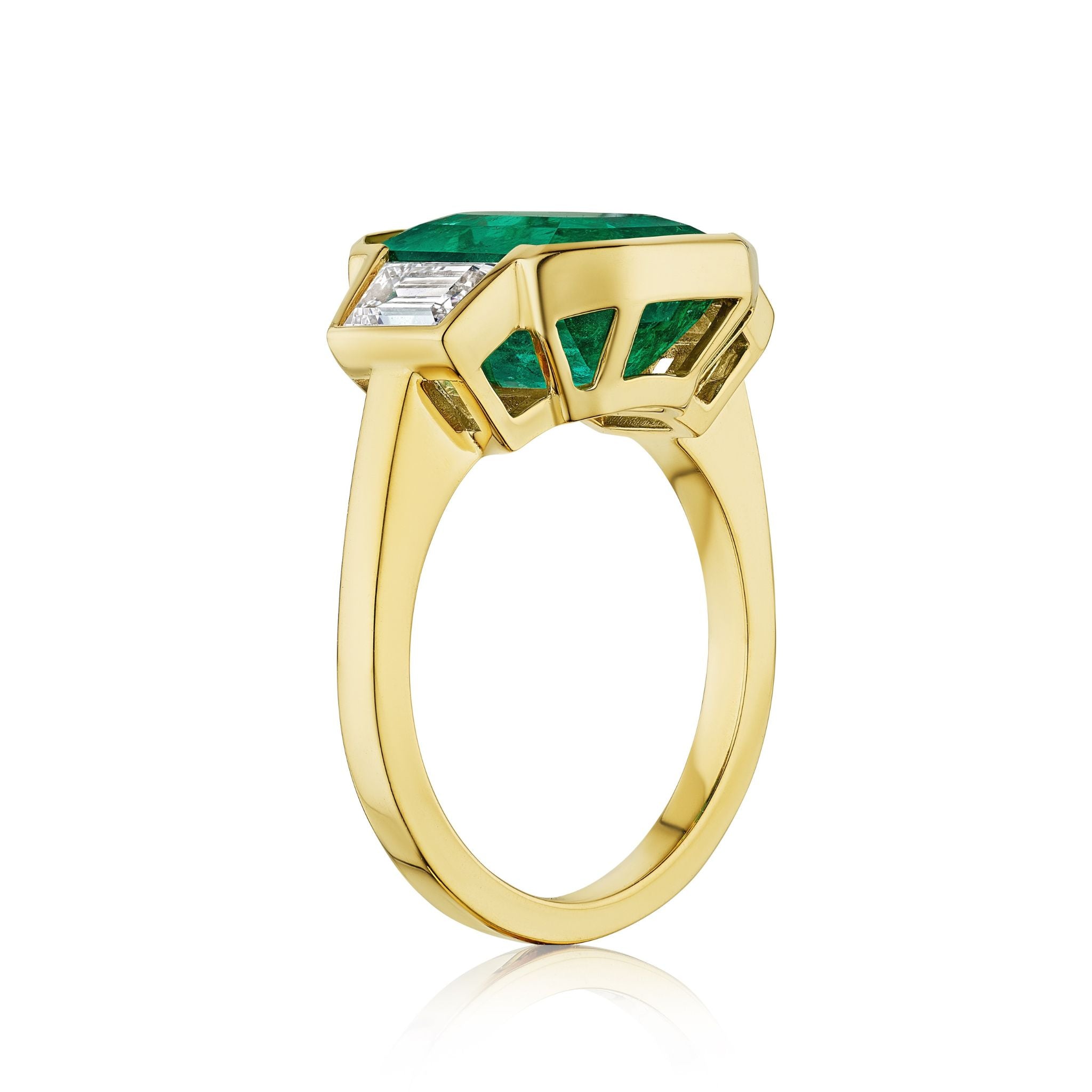 The Half Bezel Set Emerald Cut Emerald Engagement Ring features trapezoid diamond side stones and a polished 18k yellow gold band, shown at an angle to highlight its modern geometric setting.
