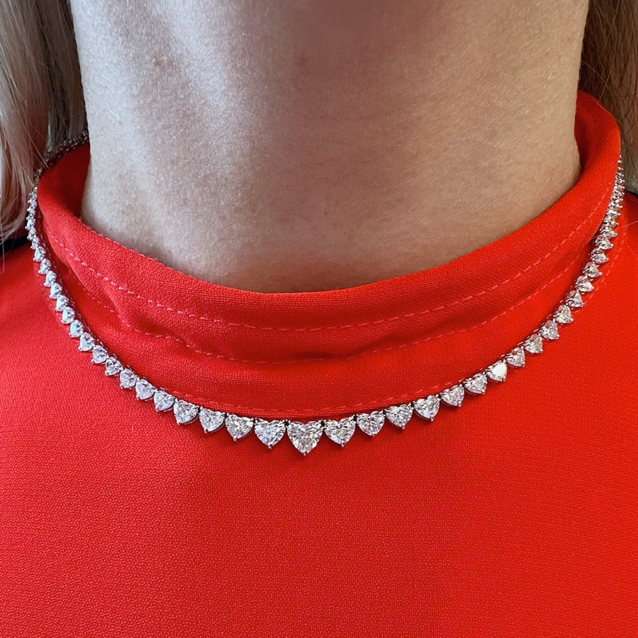 A close-up of a person's neck wearing the Graduated Heart Diamond Tennis Necklace in White Gold, featuring sparkling heart-shaped diamonds, paired with a bright red, high-collared top.