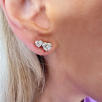 Close-up of Duo Studs with Heart Diamonds: two heart-shaped diamond studs, one larger than the other, in 18k white and rose gold, worn on a light-skinned person with blonde hair.