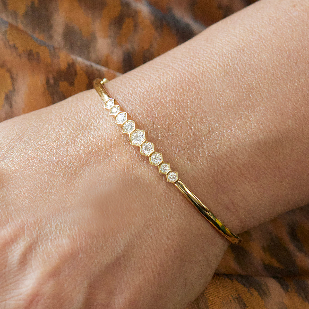 The Graduated Hexagon Bezel Set Diamond Bangle, showcasing alternating hexagon and round diamonds, sparkles on a wrist against a brown patterned fabric background.