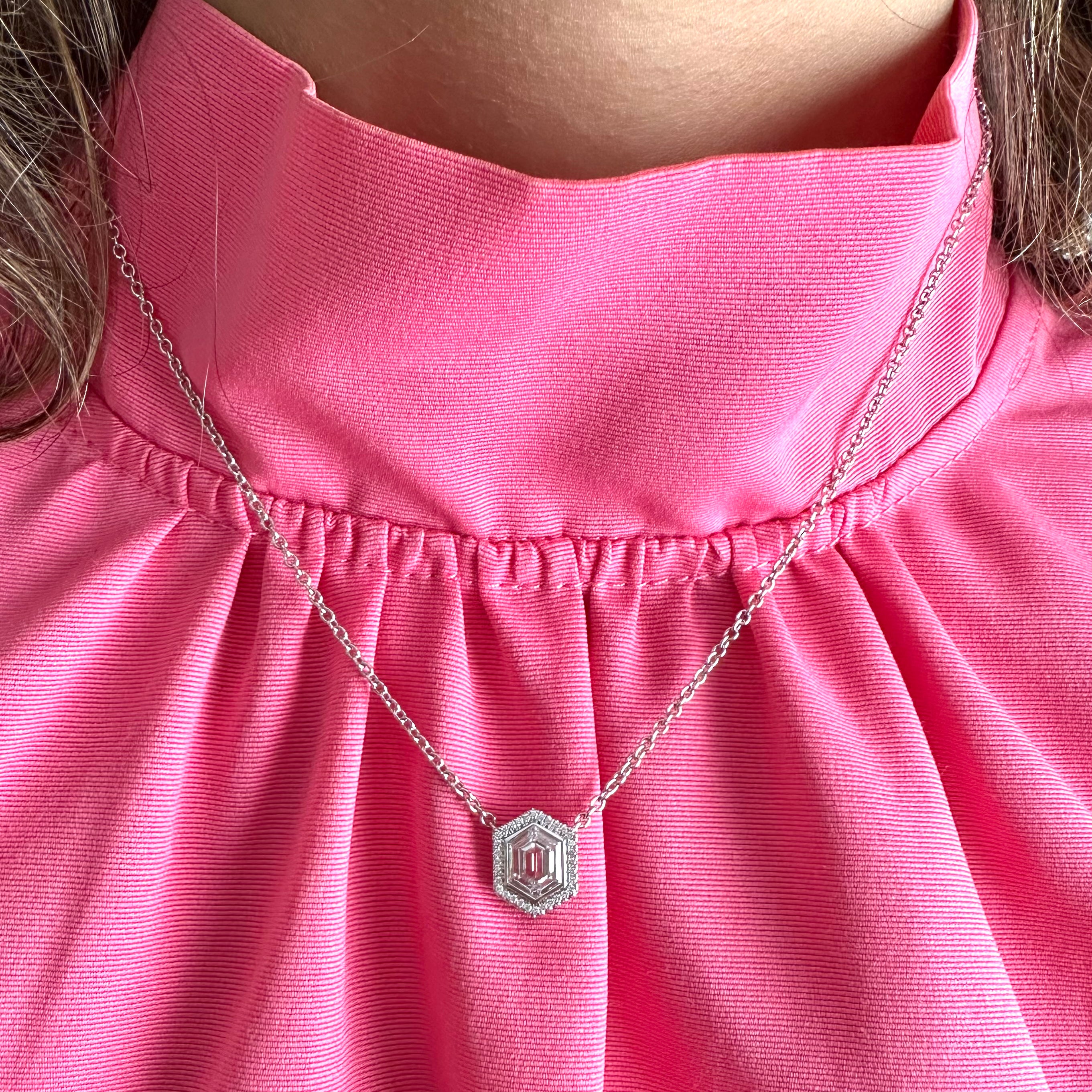 A person wears a bright pink blouse with a pleated collar, highlighting the Pavé Hexagonal Diamond Solitaire Pendant in White Gold—an elegant 18K piece featuring a striking geometric design.