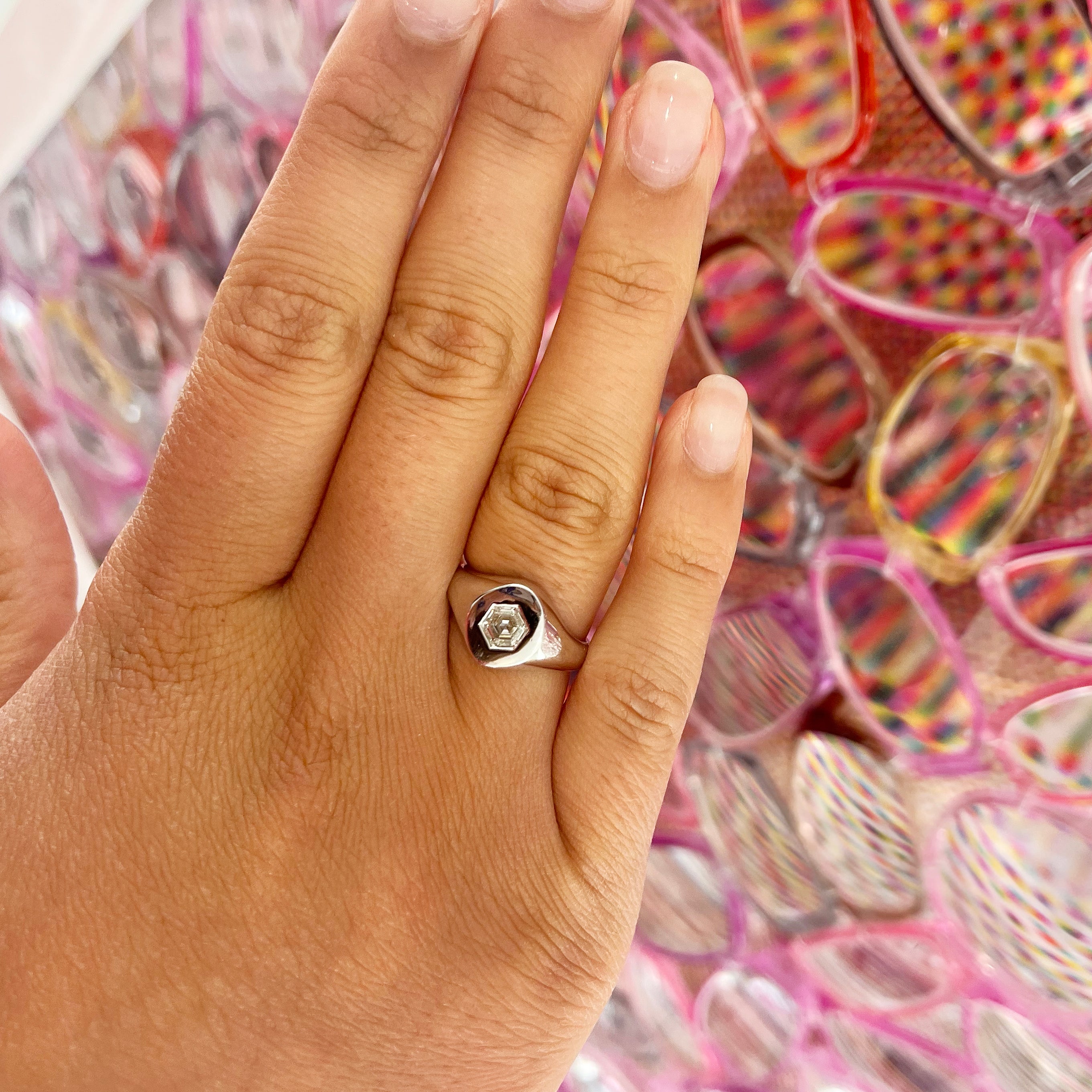 A manicured hand wears a Signet Ring with Hexagon Diamond, set against a vibrant backdrop of overlapping eyeglasses with pink and red patterned lenses.