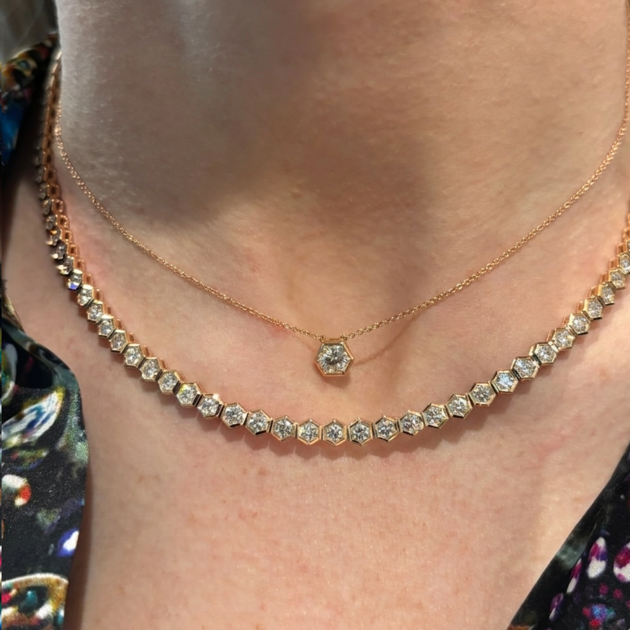 A close-up of a person wearing two layered 18K gold necklaces: one with hexagonal diamond settings and the other featuring a Hexagon Bezel Set Round Brilliant Diamond Pendant, paired with a patterned blouse.