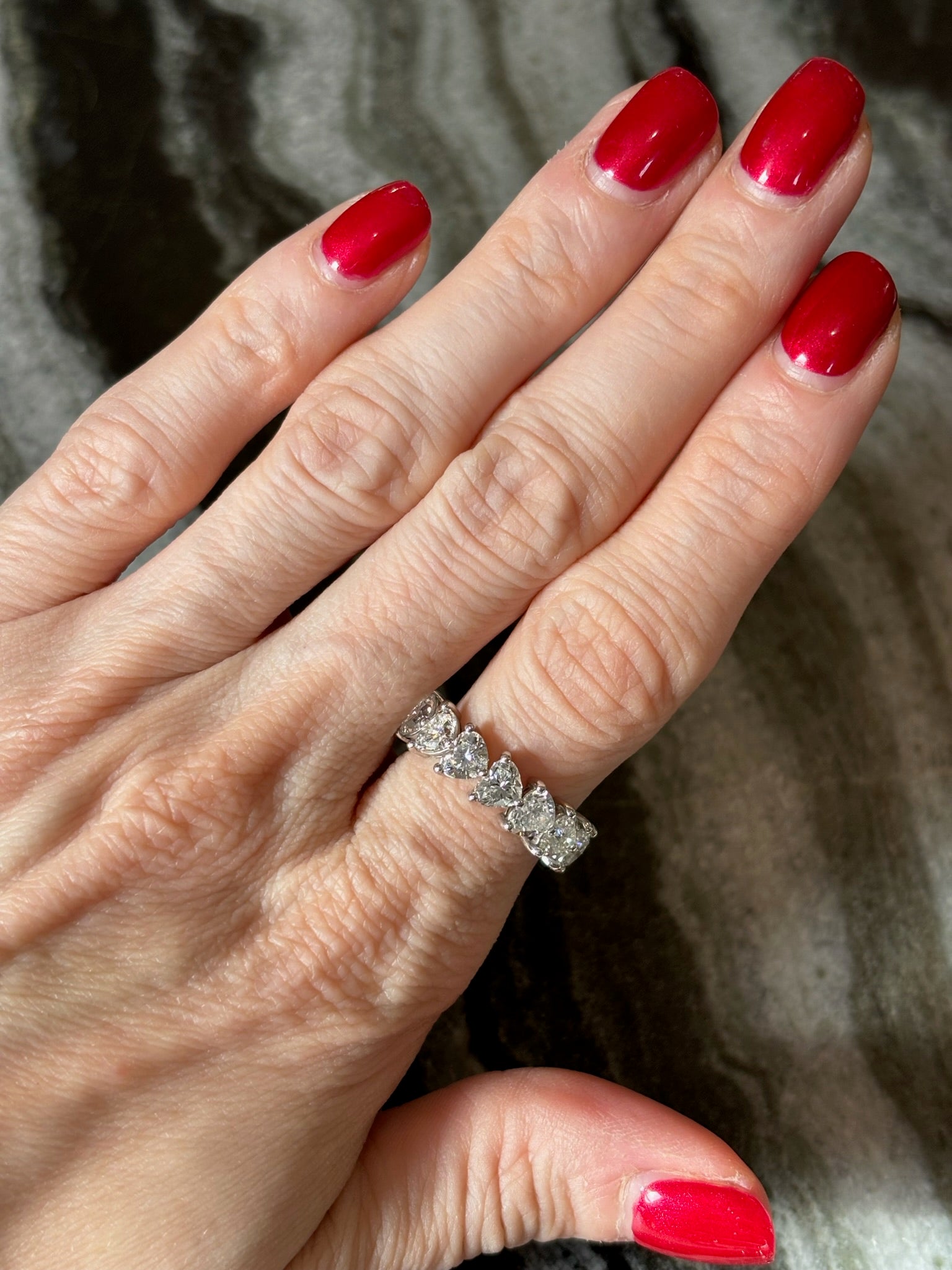 A hand with red nails displays the Chasing Hearts Eternity Band—an 18k white gold ring set with five large heart-shaped diamonds—on the ring finger, against a dark marbled background.