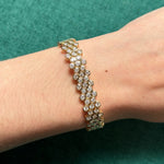 The Multi-Tier Hexagon Bezel Set Tennis Bracelet in Yellow Gold, featuring round diamonds in hexagon links, is showcased on a wrist against a green textured background.