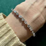 A close-up of a wrist wearing the Heart Shape Centered Mixed Shape Prong Set Diamond Tennis Bracelet in silver, featuring clear gemstones of various shapes, including a heart shape diamond, against a green background with a beige knit sweater.