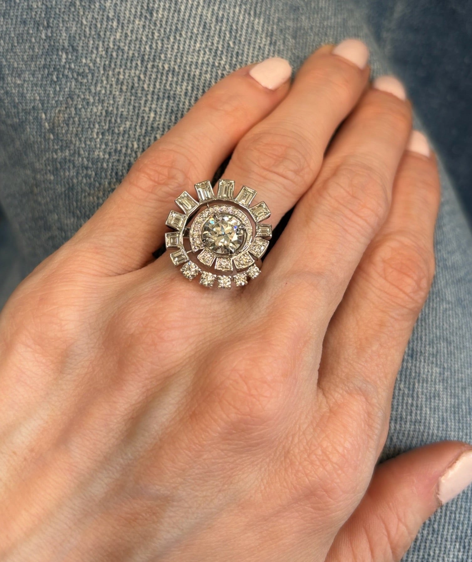 A close-up of a hand with light pink nail polish wearing the Spiral Ring in White Gold, set with round brilliant and baguette diamonds in a dazzling design, against a blue denim background.
