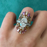 The Multicolor Tapered Baguette Sapphire and Round Diamond Ring Jacket in Yellow Gold showcases vibrant pink and yellow sapphires with sparkling diamonds—an eye-catching, customizable accessory worn on a finger against a teal background.
