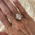 A hand with light pink nail polish wears the Oval Pink Diamond Engagement Ring with Epaulette Diamond Side Stones in Platinum and Yellow Gold, set against a cream patterned fabric background.