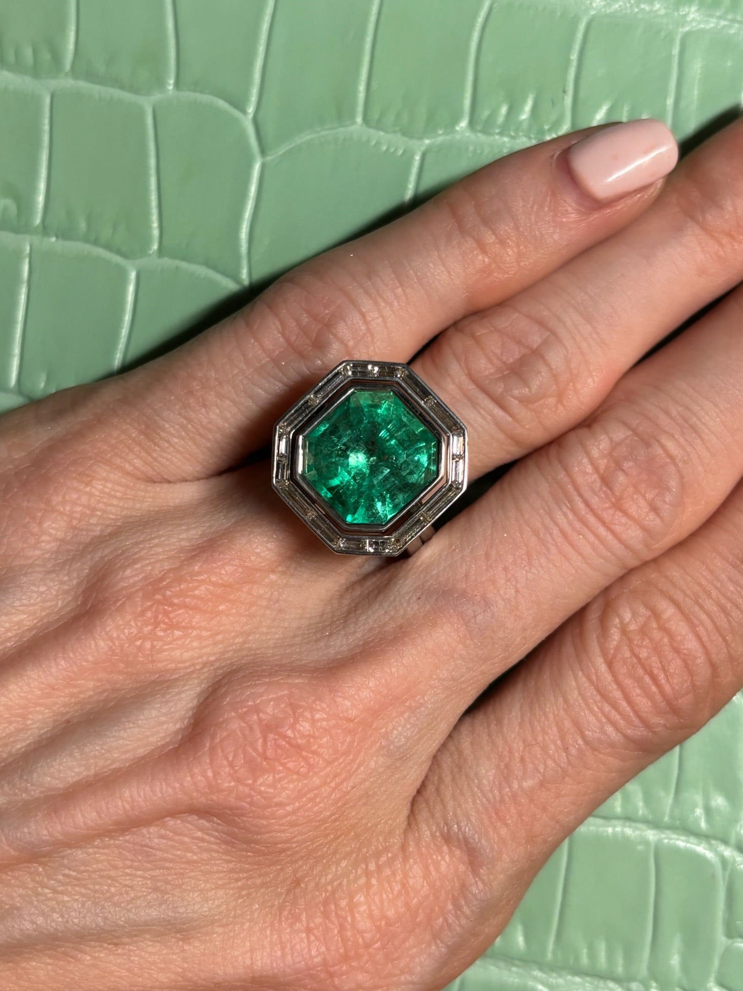 A hand with manicured nails wears a Bespoke Channel Set Trapezoid Baguette Diamond Ring Jacket in white gold, paired with a large green gemstone ring, set against a light green textured background.