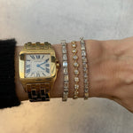 A wrist adorned with a gold and silver Cartier watch and three East-West Emerald Cut Bezel Bracelets in 18k white gold, set against a light gray background.