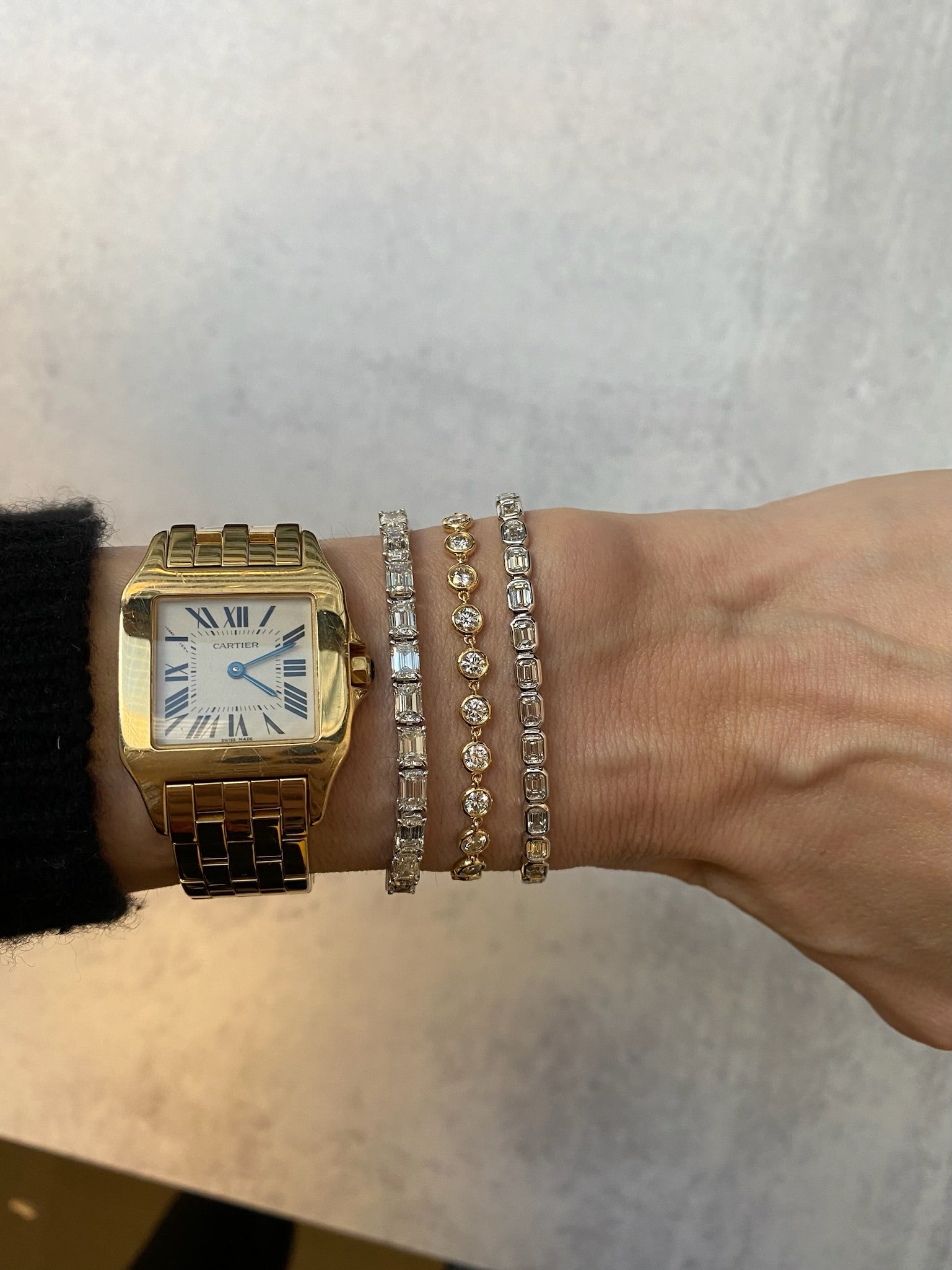 A wrist adorned with a gold and silver Cartier watch and three East-West Emerald Cut Bezel Bracelets in 18k white gold, set against a light gray background.