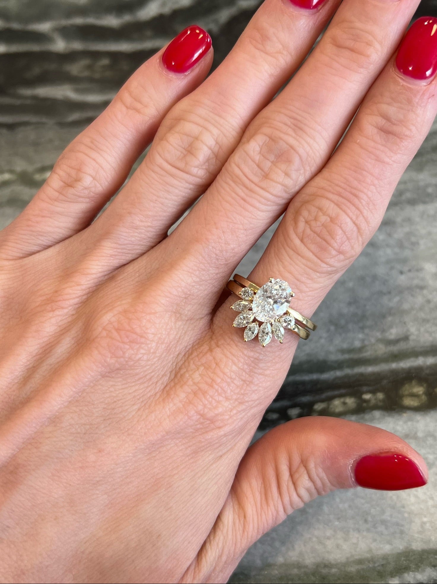A hand with red-painted nails shows two rings: a gold band and an oval diamond engagement ring accented by a Bespoke Marquise and Round Diamond Ring Jacket in yellow gold.