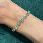 A close-up of a wrist wearing the Mixed Shape Collet Set Diamond Tennis Bracelet in white gold, featuring seven large diamonds totaling 13.38 ctw, displayed against a teal fabric background.