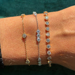 Three delicate chain bracelets, including the Milgrain Hexagon Bezel Set Garland Bracelet with round brilliant diamonds, shine in gold and silver tones on a wrist against a teal fabric background.