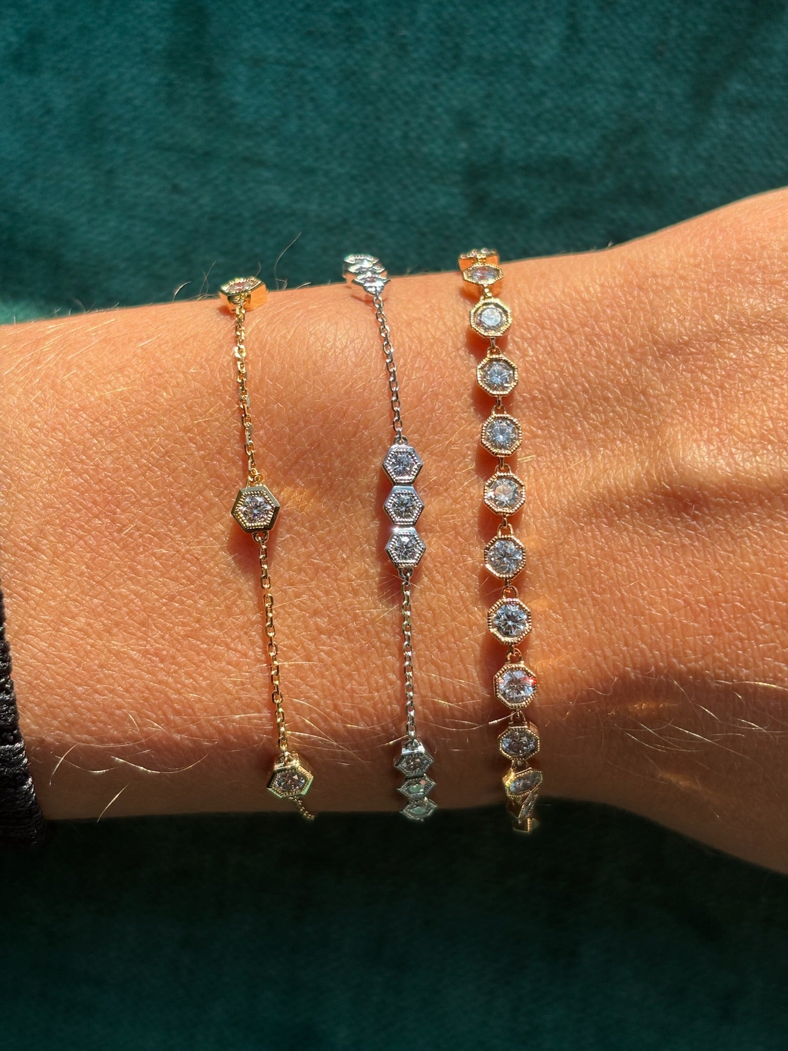 Three delicate chain bracelets, including the Milgrain Hexagon Bezel Set Garland Bracelet with round brilliant diamonds, shine in gold and silver tones on a wrist against a teal fabric background.