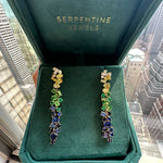 A pair of Multicolor Drop Earrings in Yellow Gold with Mixed Shape Sapphires and Diamonds, displayed in a green Serpentine Jewels box near a window with city buildings in the background.