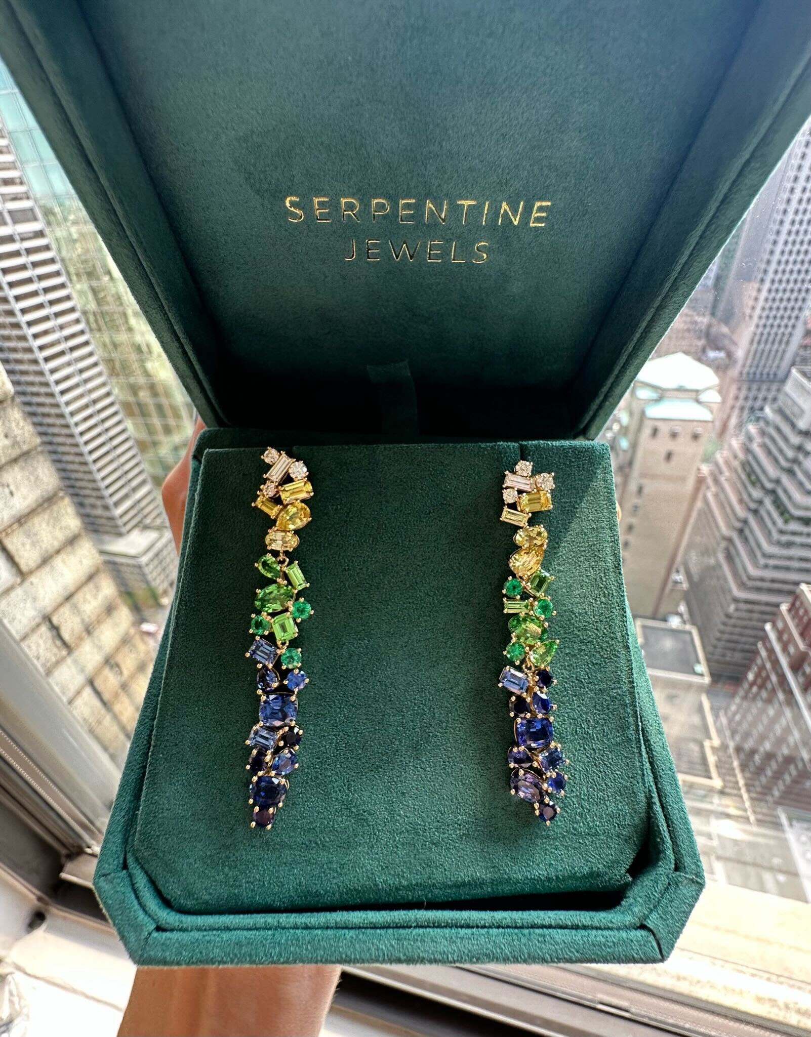 A pair of Multicolor Drop Earrings in Yellow Gold with Mixed Shape Sapphires and Diamonds, displayed in a green Serpentine Jewels box near a window with city buildings in the background.