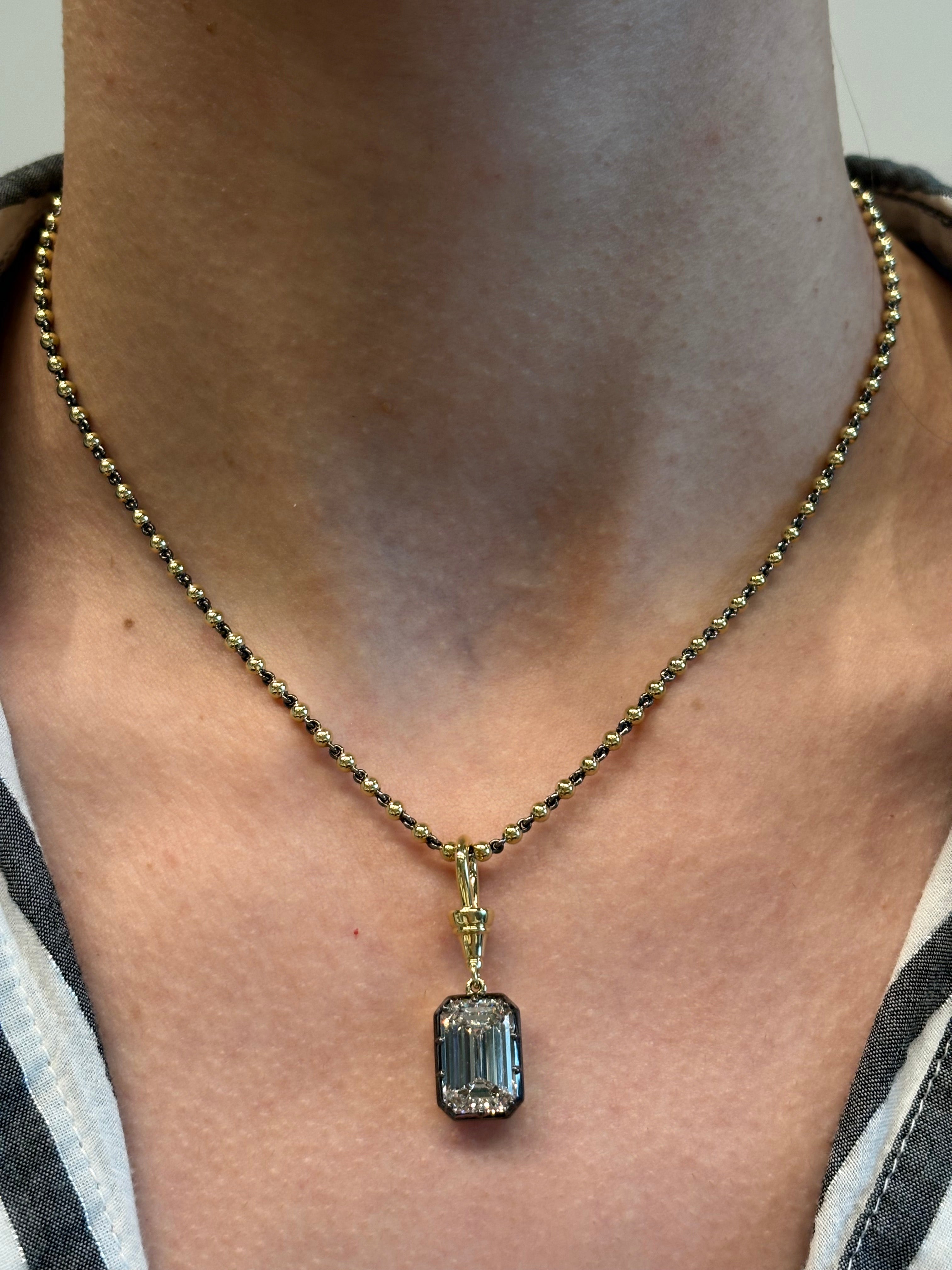 A close-up of a person wearing the Collet Set Emerald Cut Diamond in Black Rhodium and Yellow Gold, featuring an emerald cut diamond pendant on bare skin with part of a striped shirt visible.