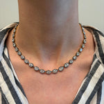 A close-up of a person's neck adorned with the Collet Set Oval Diamond Tennis Necklace in Black Rhodium. The 18k gold piece sparkles with round diamonds totaling 28.68ctw, worn over a black and white vertical striped collared shirt.