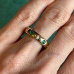 A hand wears the Burnish Set Baguette Diamond and Round Emerald Band in Yellow Gold, featuring two round green emeralds and a rectangular baguette diamond, against a green background.