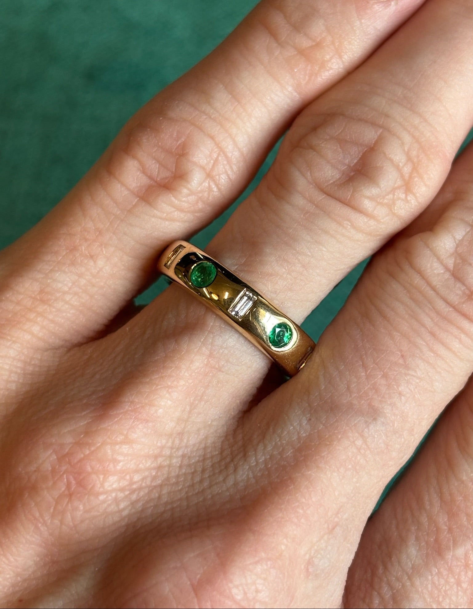 The Bespoke Burnish Set Baguette Diamond and Round Emerald Band in Yellow Gold is shown worn on a hand, featuring two round emeralds and two baguette diamonds against a green background.