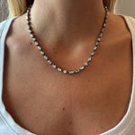 A close-up of a woman wearing a white top and the Mixed Shape Small Collet Set Diamond Necklace in Black Rhodium, featuring evenly spaced small round diamonds against her bare neck and collarbone.