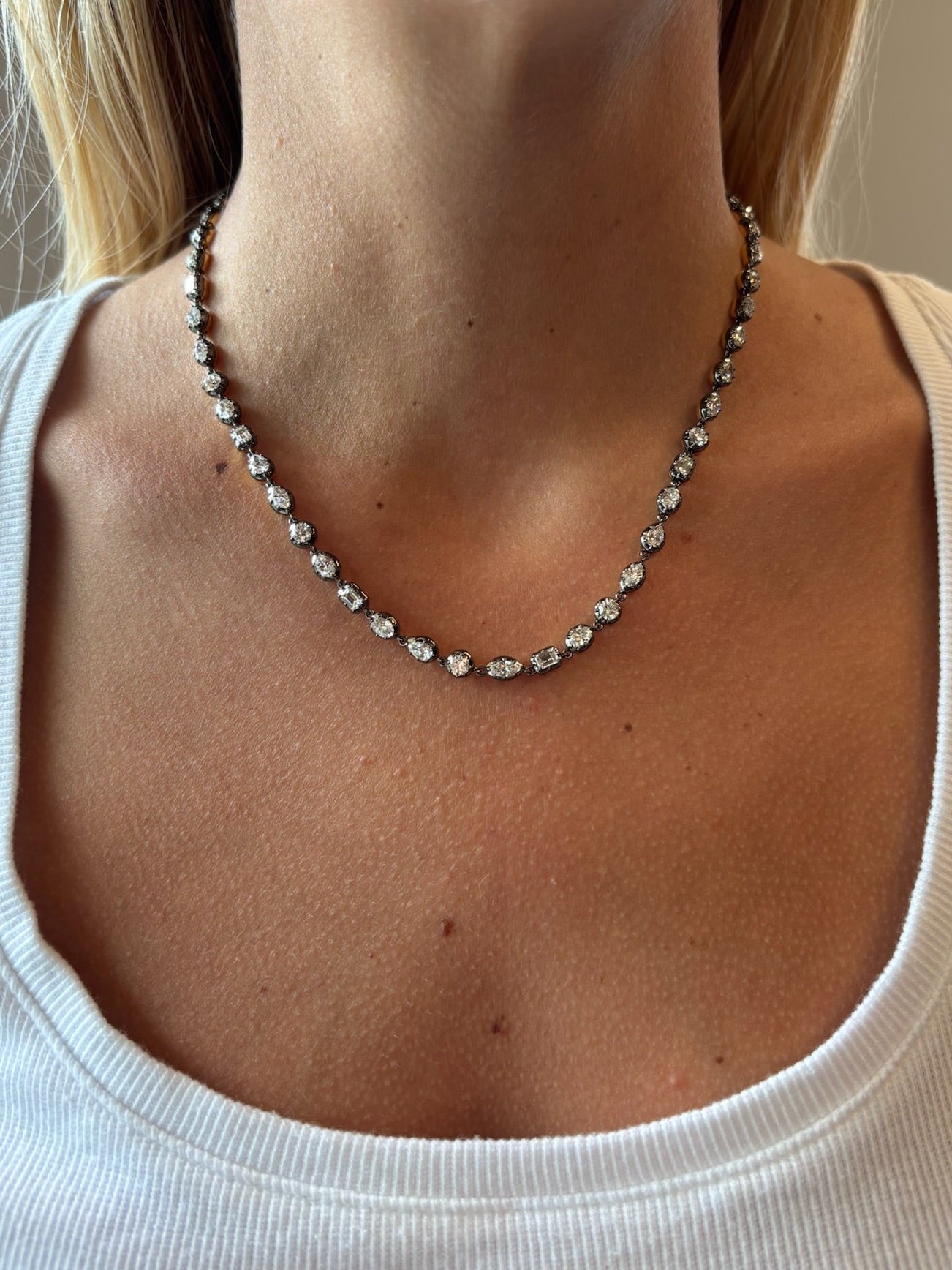 A close-up of a woman wearing a white top and the Mixed Shape Small Collet Set Diamond Necklace in Black Rhodium, featuring evenly spaced small round diamonds against her bare neck and collarbone.