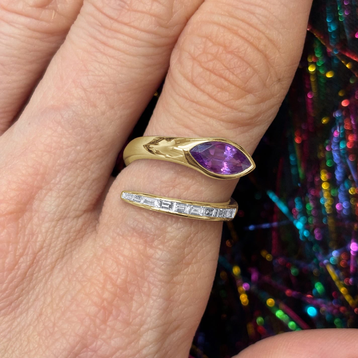 A hand with red nail polish wears the Snake Wraparound Ring with Marquise Sapphire and Baguette Diamonds in yellow gold, displayed against a colorful, shiny background.