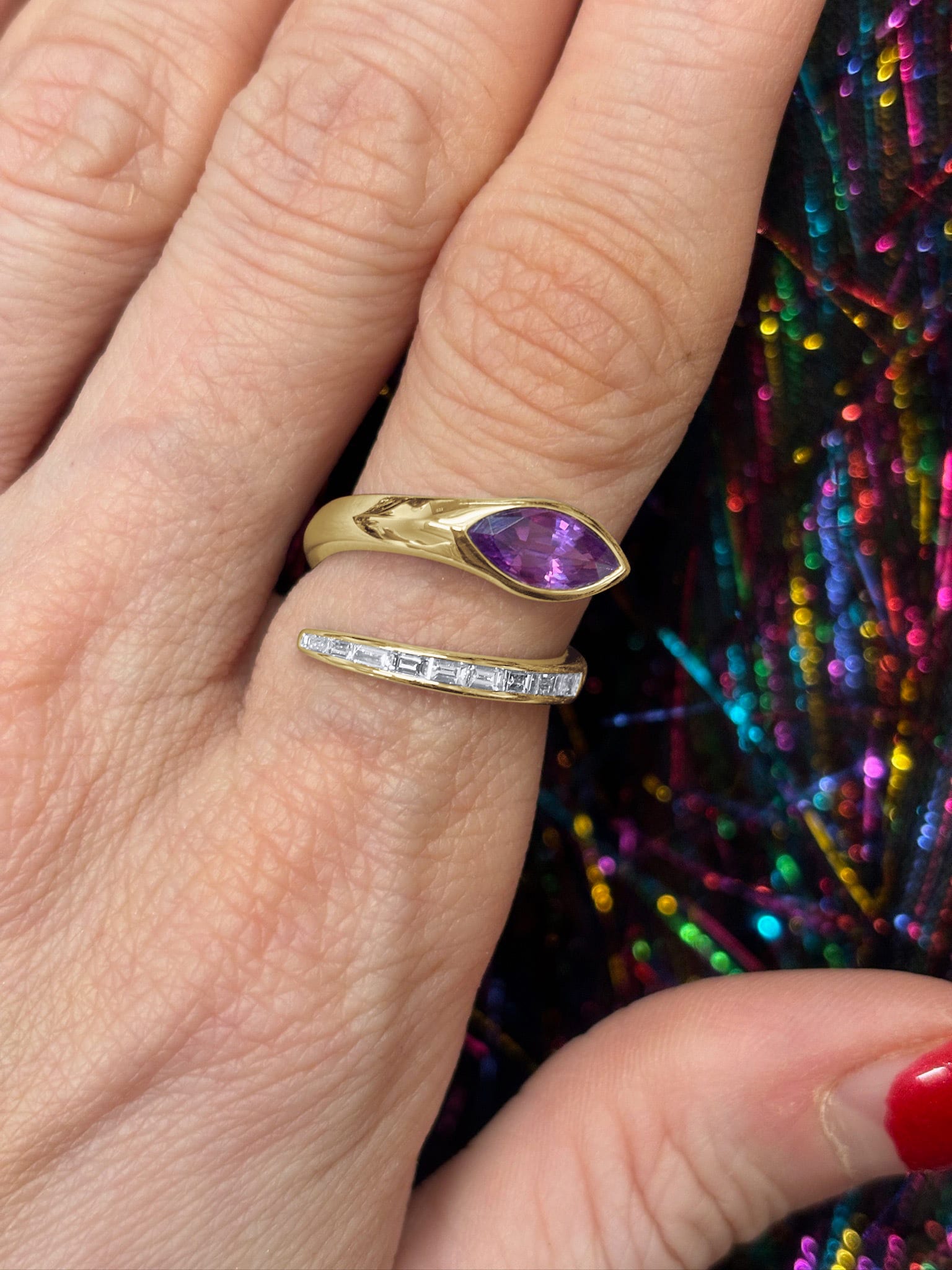 A hand with red nail polish wears the Snake Wraparound Ring with Marquise Sapphire and Baguette Diamonds in yellow gold, displayed against a colorful, shiny background.