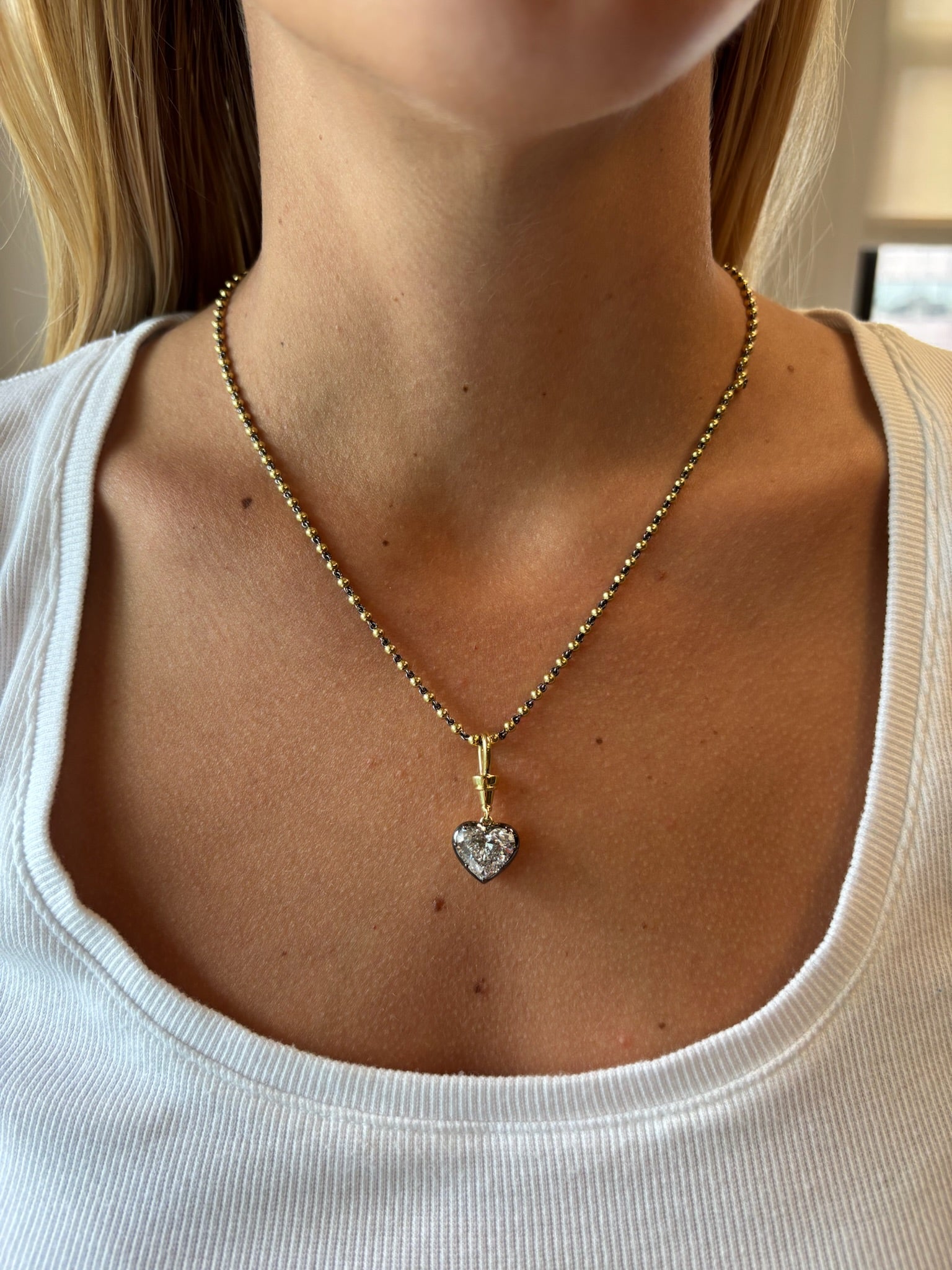 A close-up of a woman wearing the Collet Set Heart Shape Diamond Necklace in Black Rhodium and Yellow Gold against her bare neck and a white ribbed shirt.