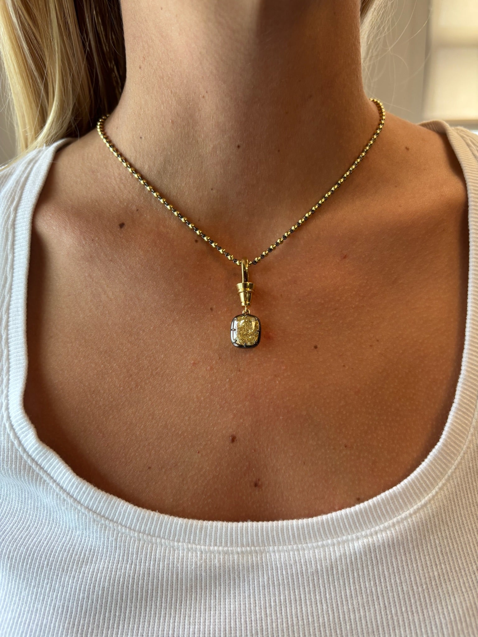 A close-up of a person wearing the Collet Set Cushion Cut Yellow Diamond Necklace in Black Rhodium and Yellow Gold with a white scoop-neck top, highlighting the jewelry and collarbone area.