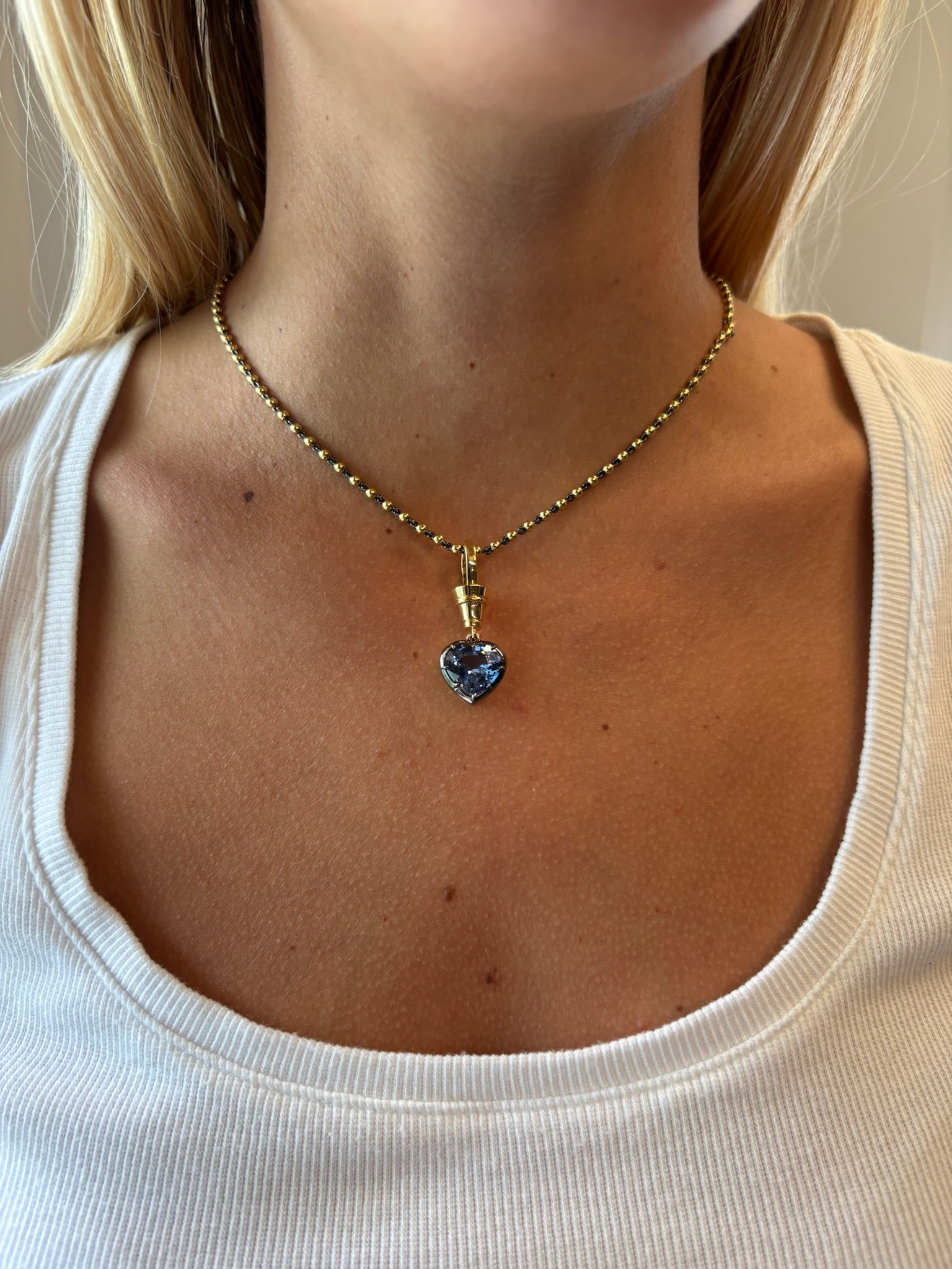 A woman wears a white top and the Collet Set Heart Shape Blue Sapphire Necklace in Black Rhodium and Yellow Gold, with focus on her neck and collarbone.