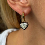 A close-up of a person with light skin and blond hair wearing the Collet Set Heart Shape Earring in Black Rhodium and Yellow Gold, featuring a gold hoop with a heart-shaped charm adorned with a clear, faceted gem at its center.