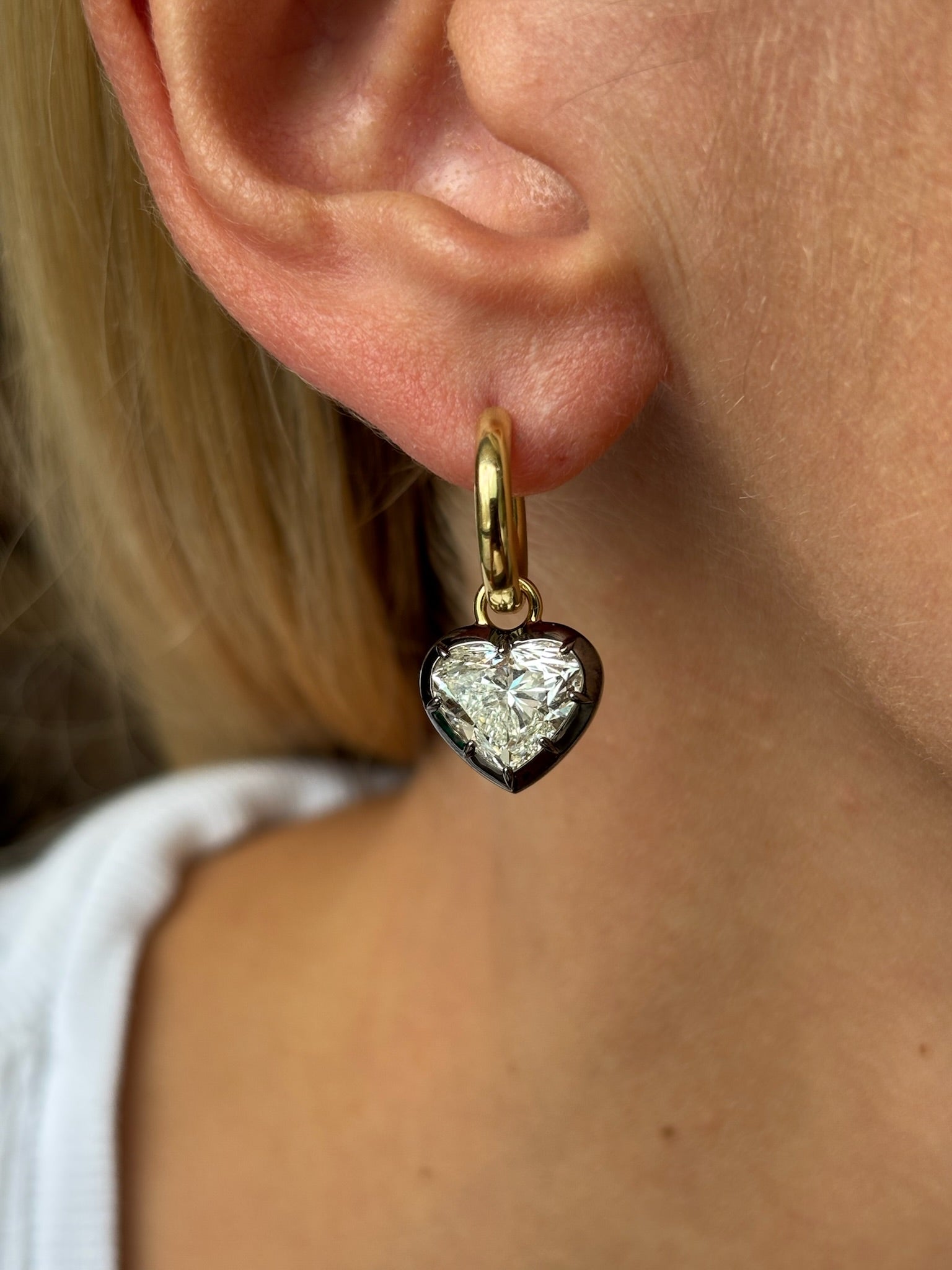 A close-up of a person with light skin and blond hair wearing the Collet Set Heart Shape Earring in Black Rhodium and Yellow Gold, featuring a gold hoop with a heart-shaped charm adorned with a clear, faceted gem at its center.