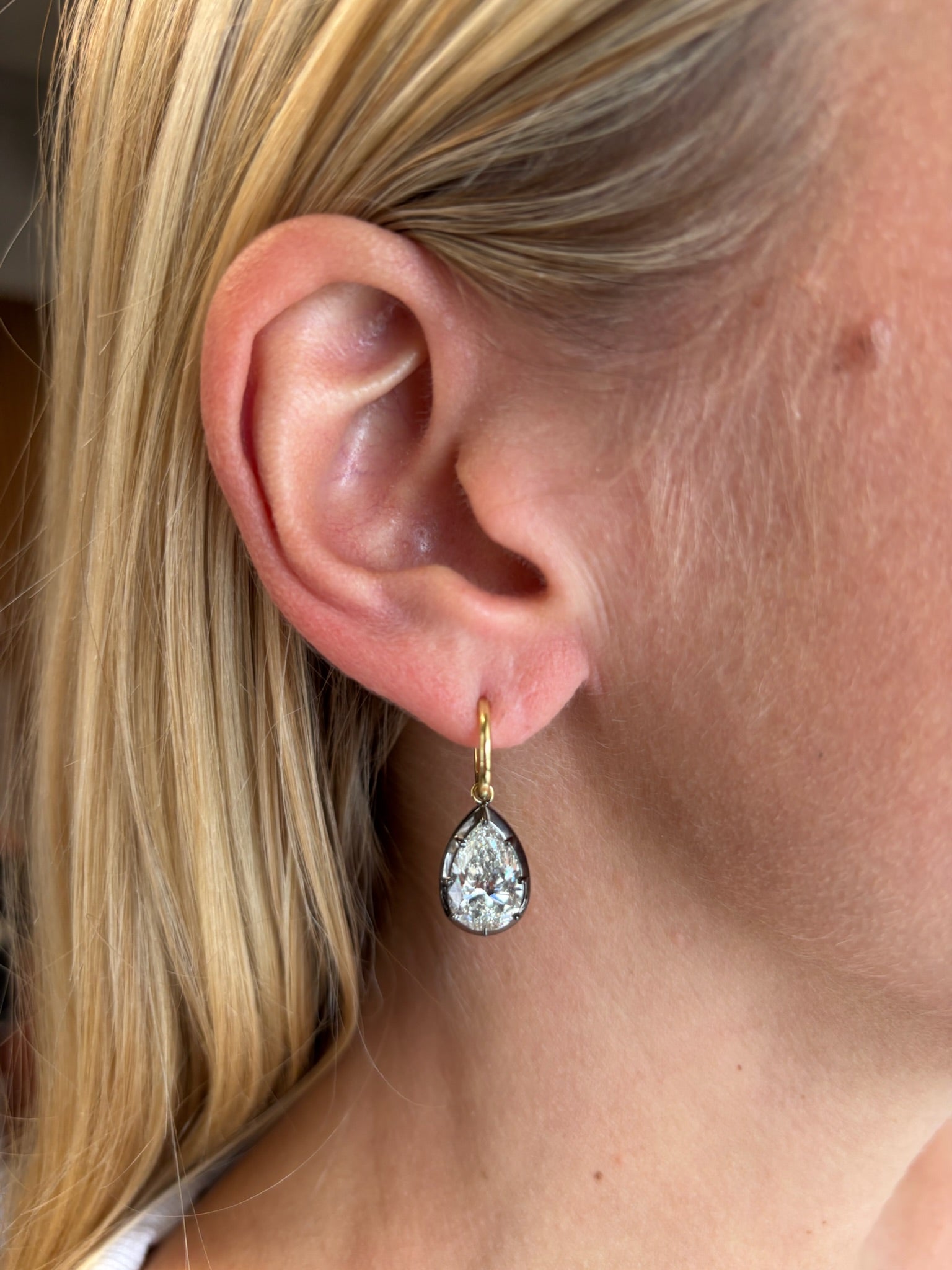 Close-up of a woman's ear with blonde hair tucked behind, showcasing the Collet Set Pear Shape Diamond Drop Earring in Black Rhodium and Yellow Gold featuring a large teardrop diamond pendant.
