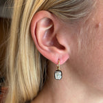 Close-up of an ear with straight blonde hair wearing the Collet Set Cushion Cut Diamond Earring in Black Rhodium and Yellow Gold, featuring a gold hoop and a cushion-cut diamond pendant.