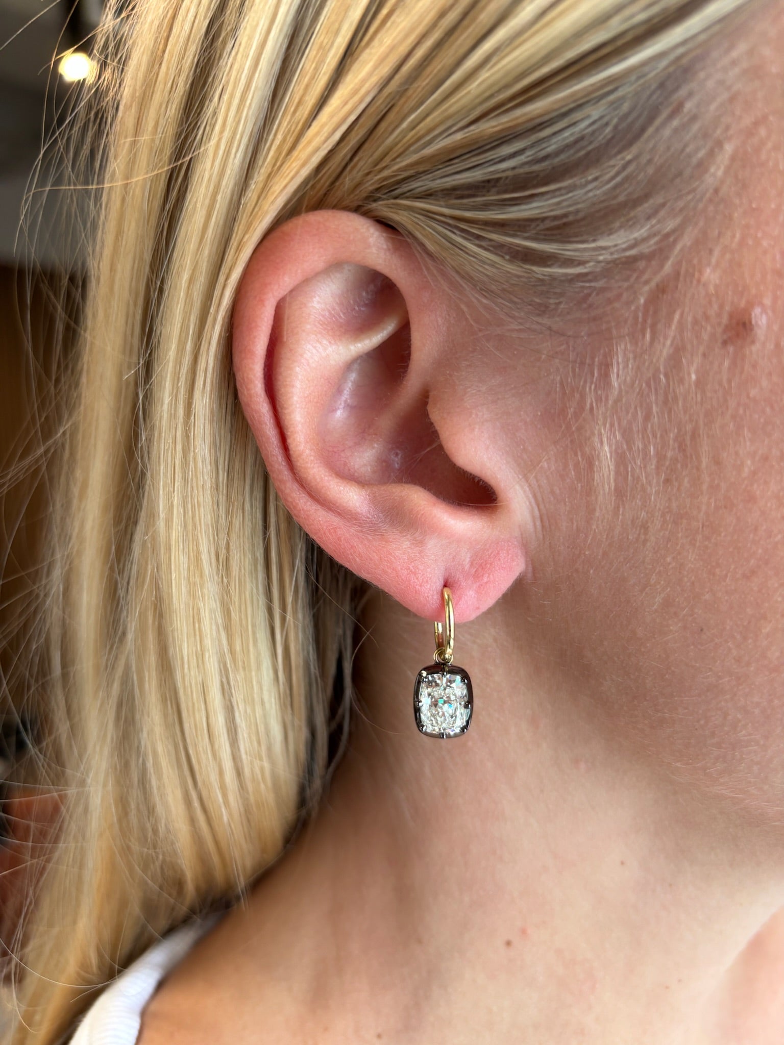 Close-up of an ear with straight blonde hair wearing the Collet Set Cushion Cut Diamond Earring in Black Rhodium and Yellow Gold, featuring a gold hoop and a cushion-cut diamond pendant.